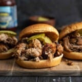 Three pulled pork sandwiches with pickles on a wooden cutting board.
