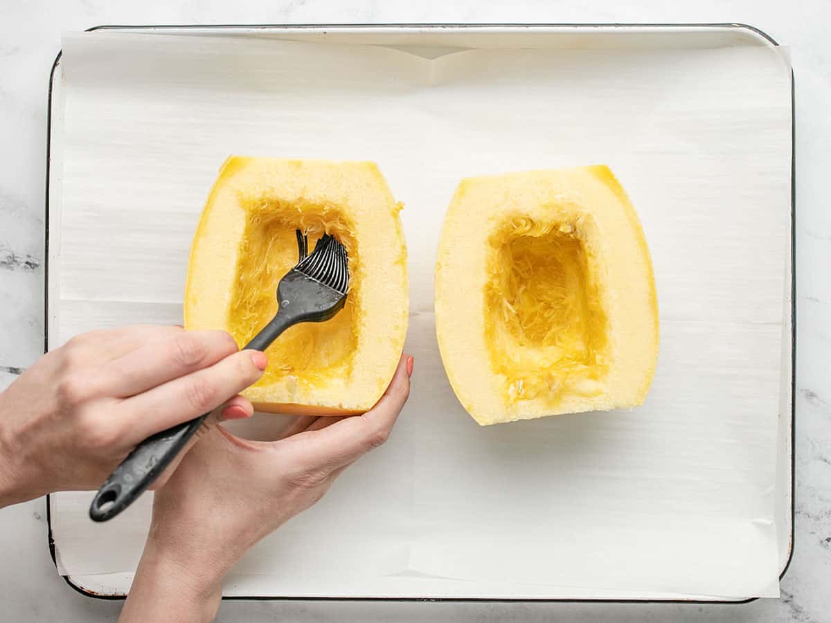 Overhead shot of a hand holding a spaghetti squash half and brushing oil on it.