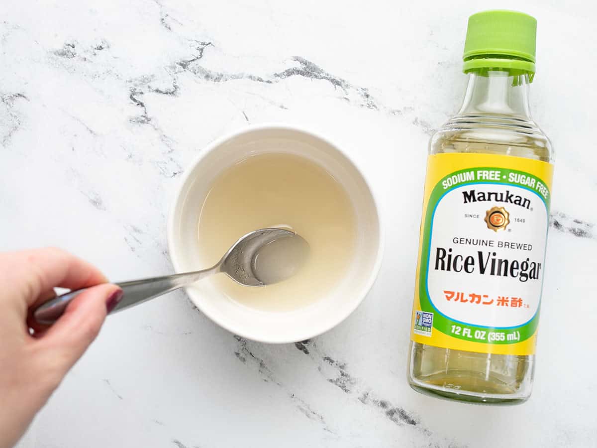 Rice vinegar, sugar, and salt being stirred in a small bowl, the bottle of vinegar on the side.