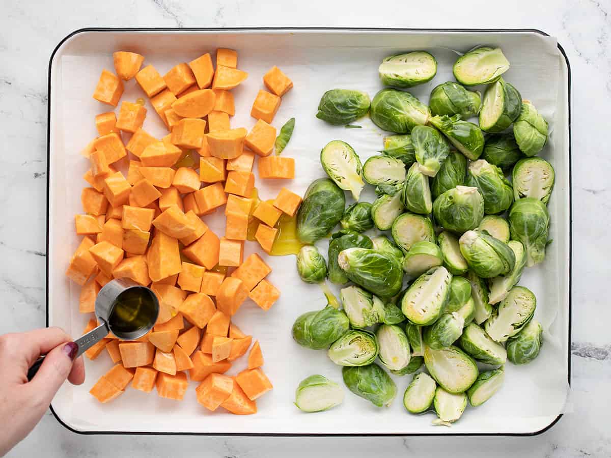 Brussels sprouts and sweet potatoes being seasoned on a sheet pan.