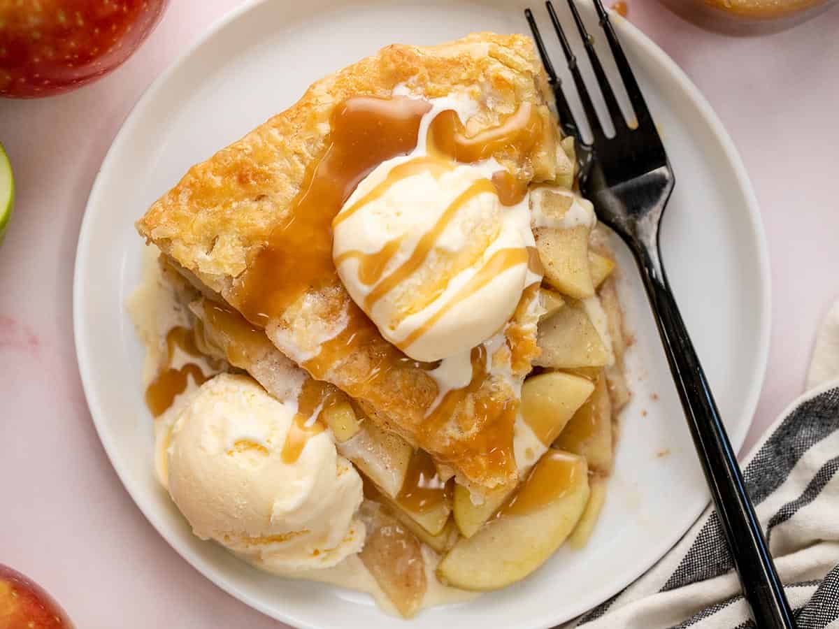 Overhead shot of a slice of apple pie on a white plate with two scoops of vanilla ice cream and drizzled with caramel sauce.