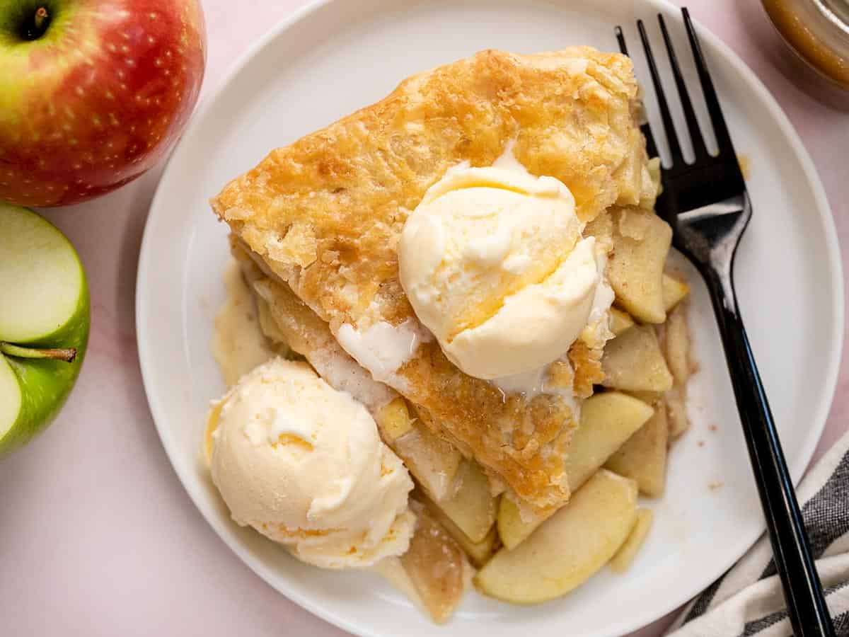 Overhead shot of a slice of apple pie on a white plate with two scoops of vanilla ice cream and drizzled with caramel sauce.