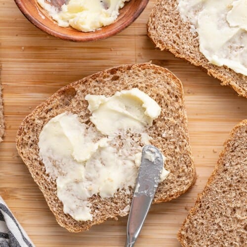 Overhead view of butter being spread on slices of wheat bread.