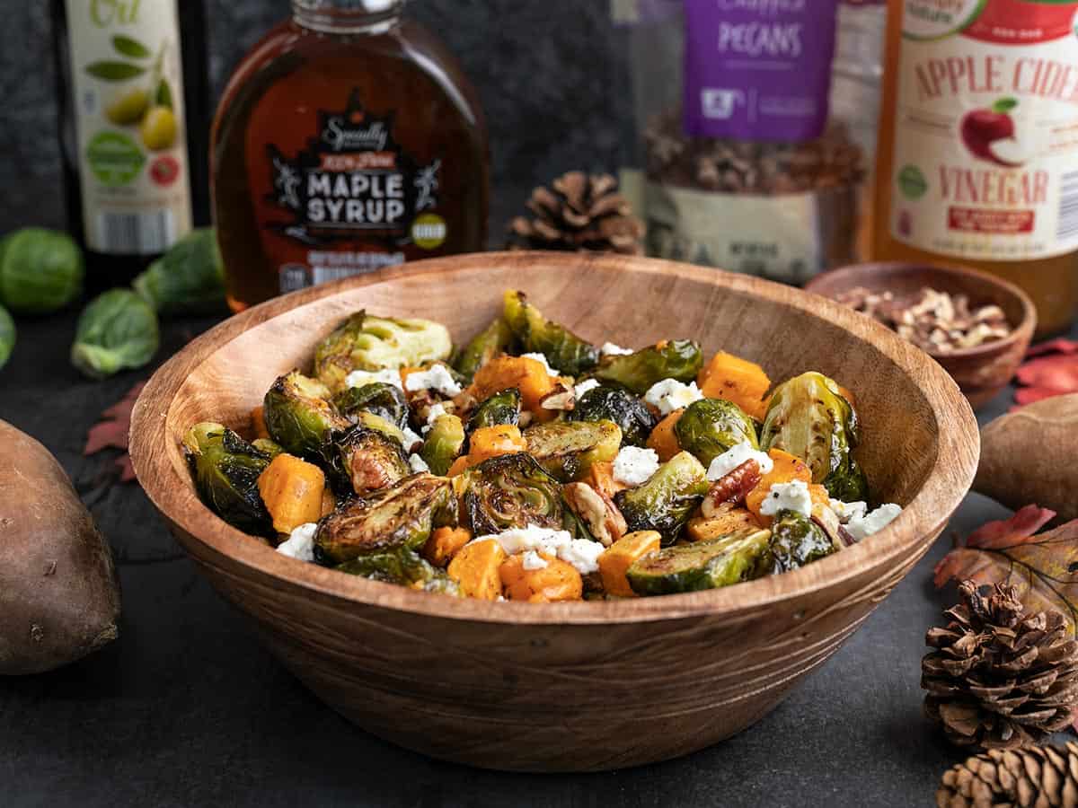 Side view of a wooden bowl full of roasted brussels sprouts salad with ingredients in the back.