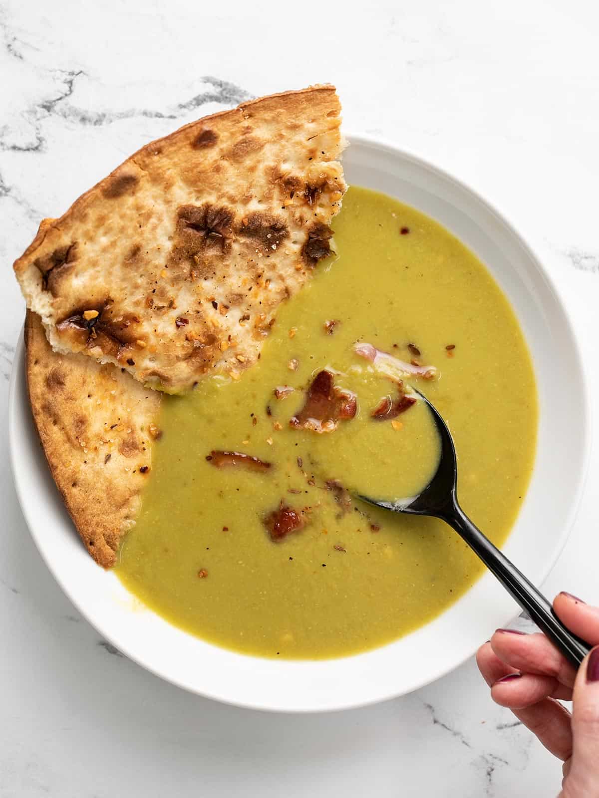 Overhead view of a bowl of split pea soup with flatbread and a spoon dipping into the center.