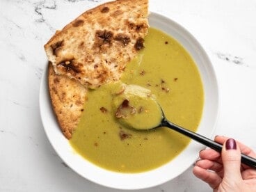 A spoon dipping into a bowl of pea soup with flatbread on the side.