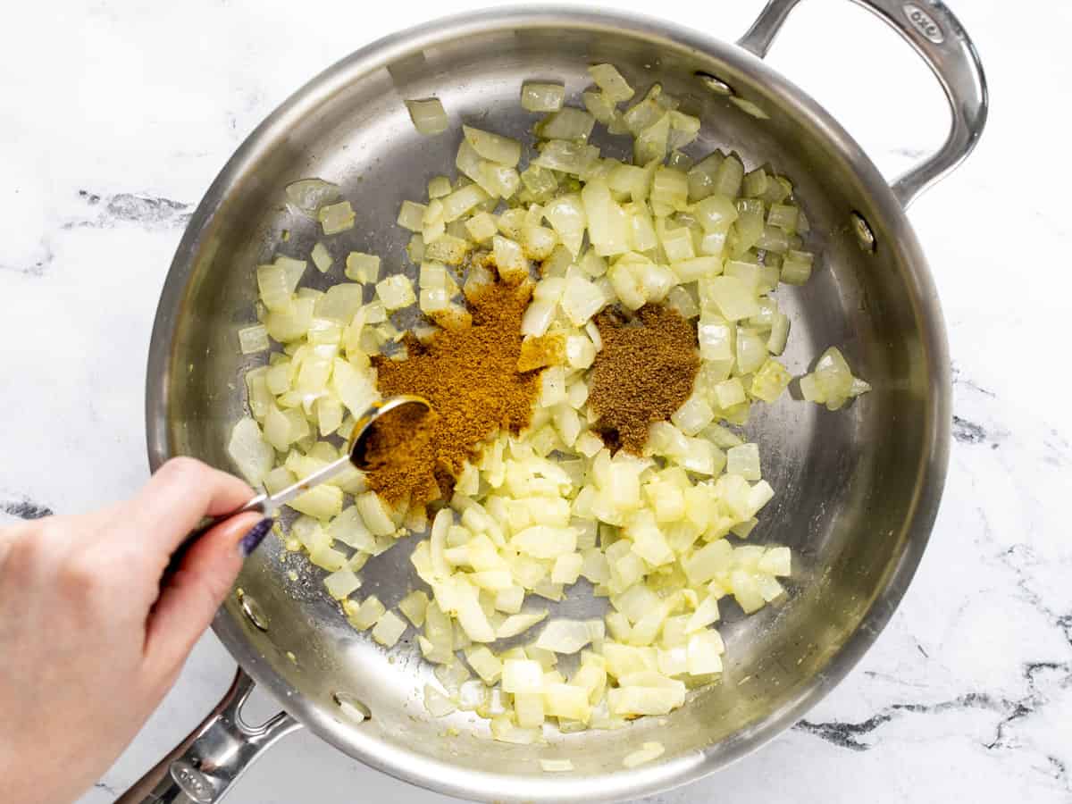 Sautéed aromatics in a skillet, spices being added. 