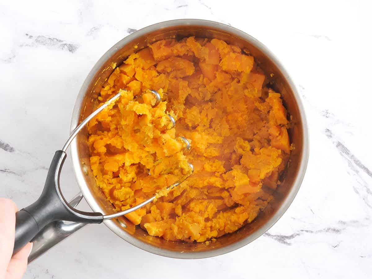 Mashed sweet potatoes in a sauce pan with a masher.