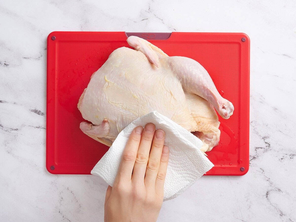 A hand using a paper towel to pat a whole chicken dry.