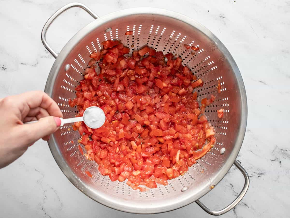 Overhead shot of a hand salting tomatoes in a silver colander.