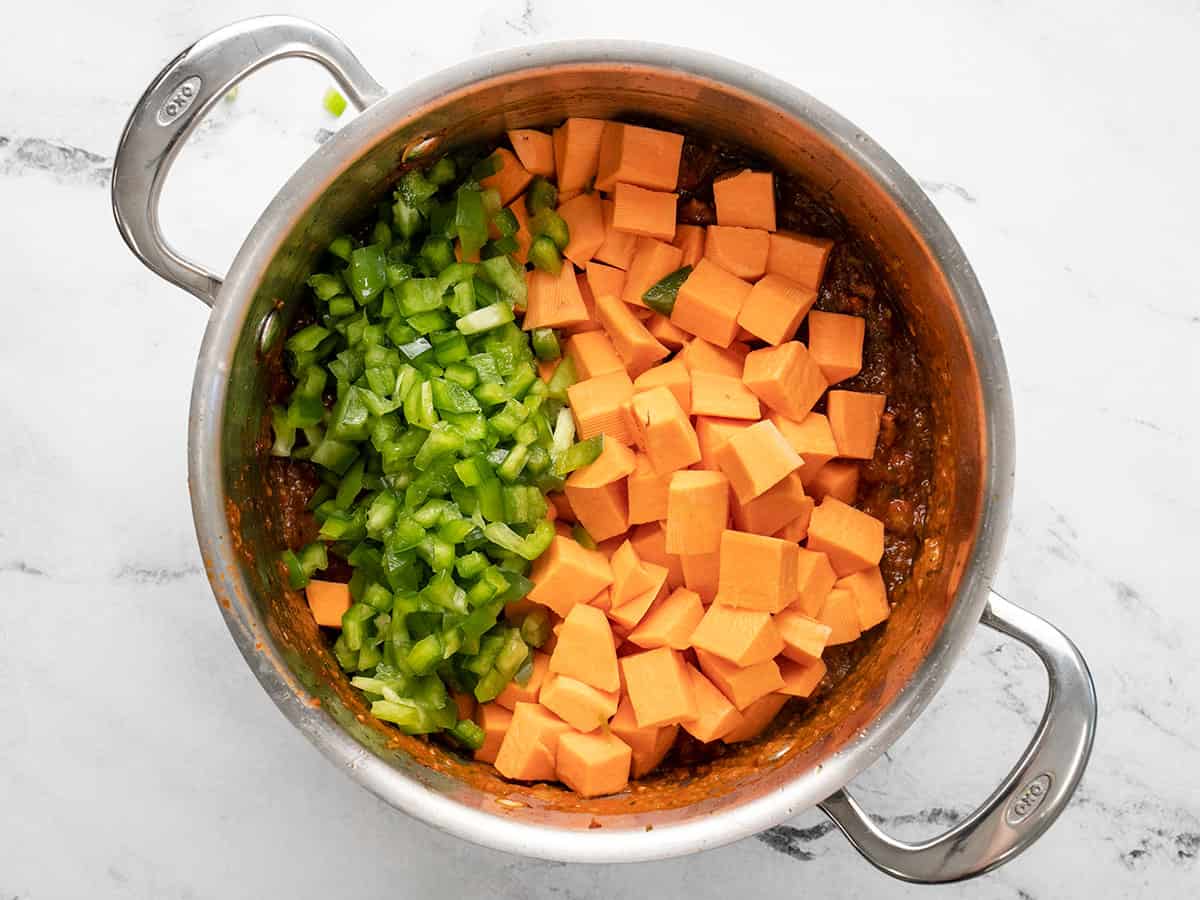 Overhead shot of diced green bell peppers and diced sweet potatoes in a silver pot with sofrito. 