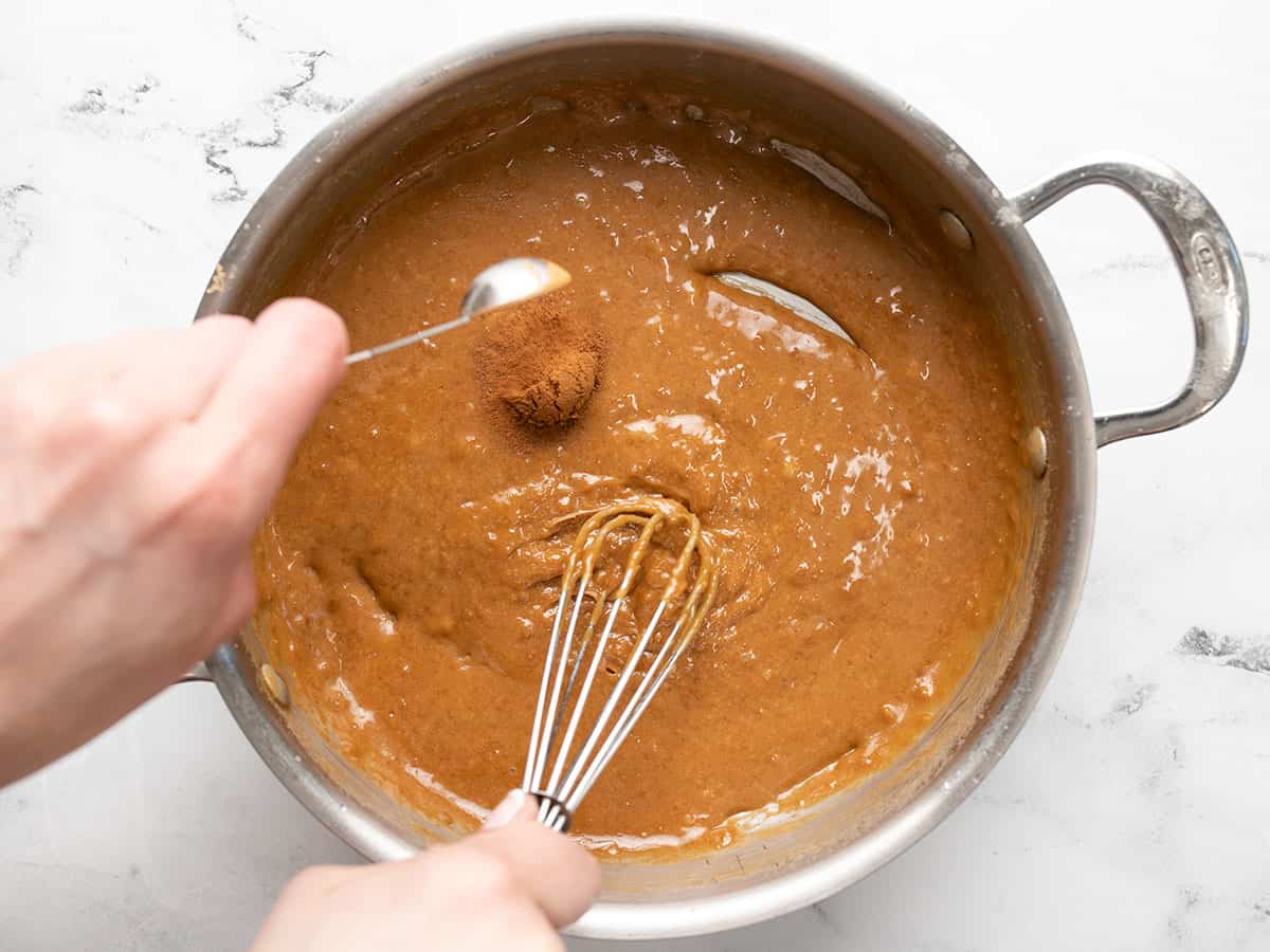 Overhead shot of hand stirring caramel with a whisk in a pot and a second hand adding vanilla to the pot with a measuring spoon.