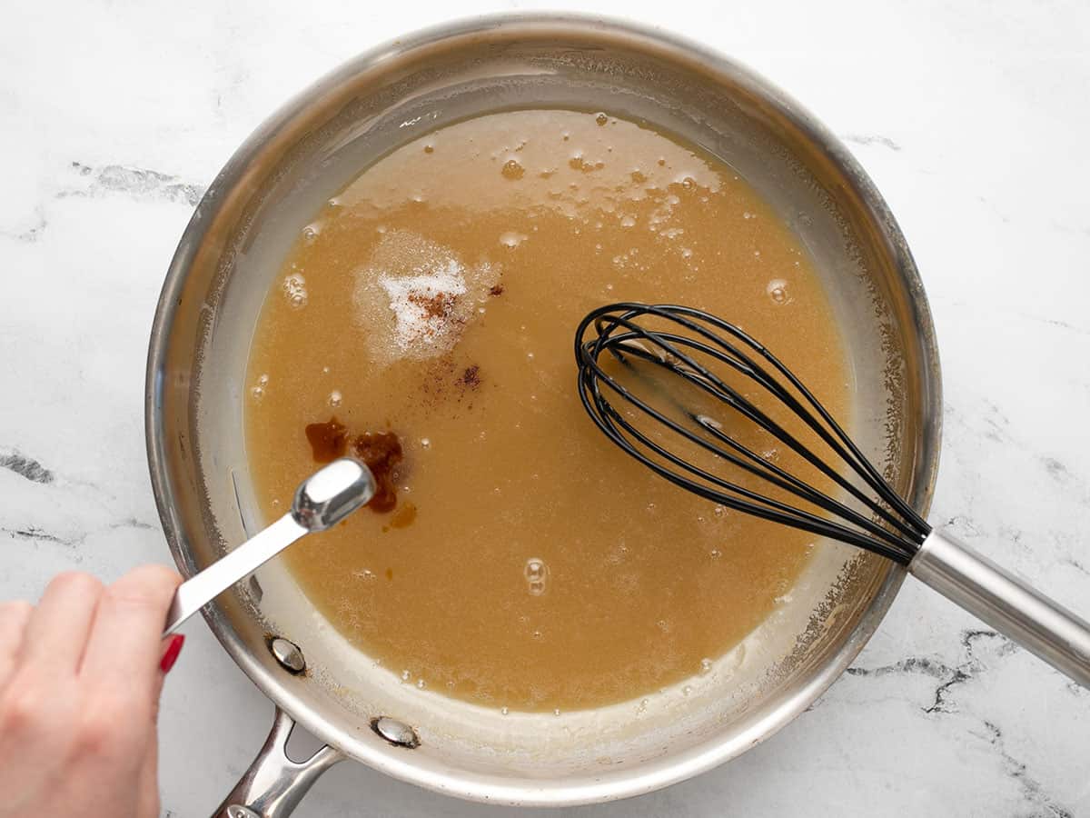Overhead shot of a spoonful of cinnamon being added to caramel in a silver pan with a whisk in it.