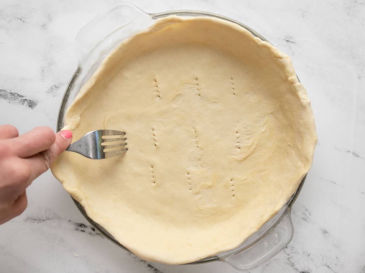 Pie crust in a pie dish being pricked with a fork. 
