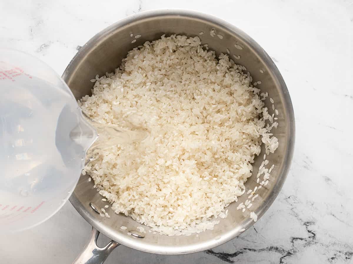 Overhead shot of water being poured into toasted rice in a silver pot.