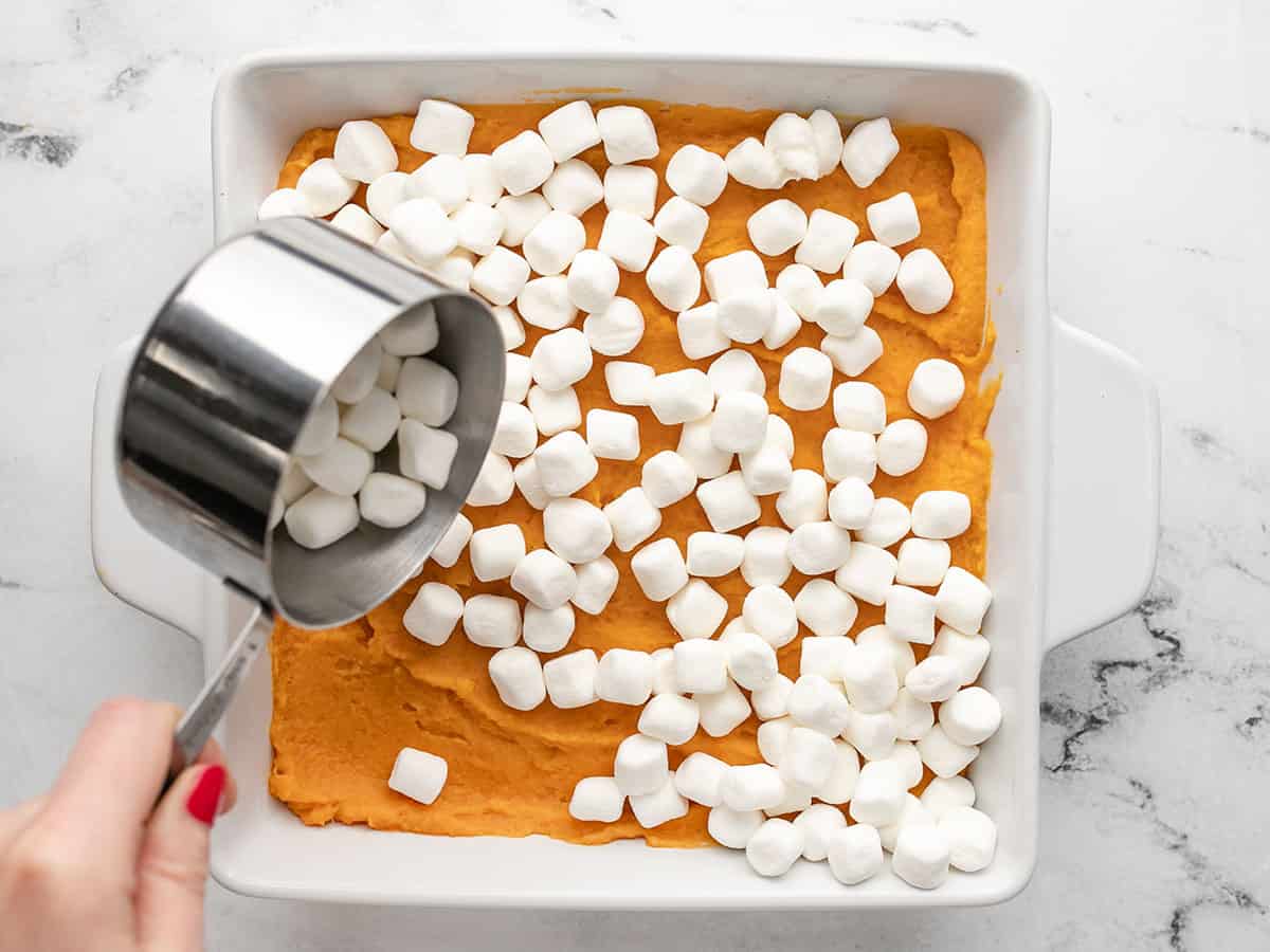 Overhead shot of a silver cup measure pouring a cup of marshmallows into a sweet potato casserole with marshmallows already on top of it.