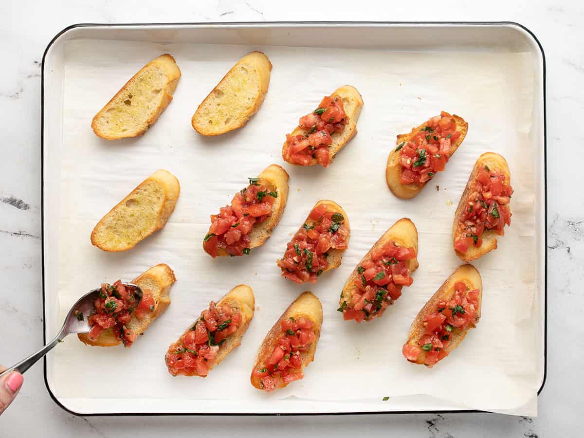 Overhead shot of a sheet pan with 13 baguette slices, some topped with chopped tomato mixture and others bare.