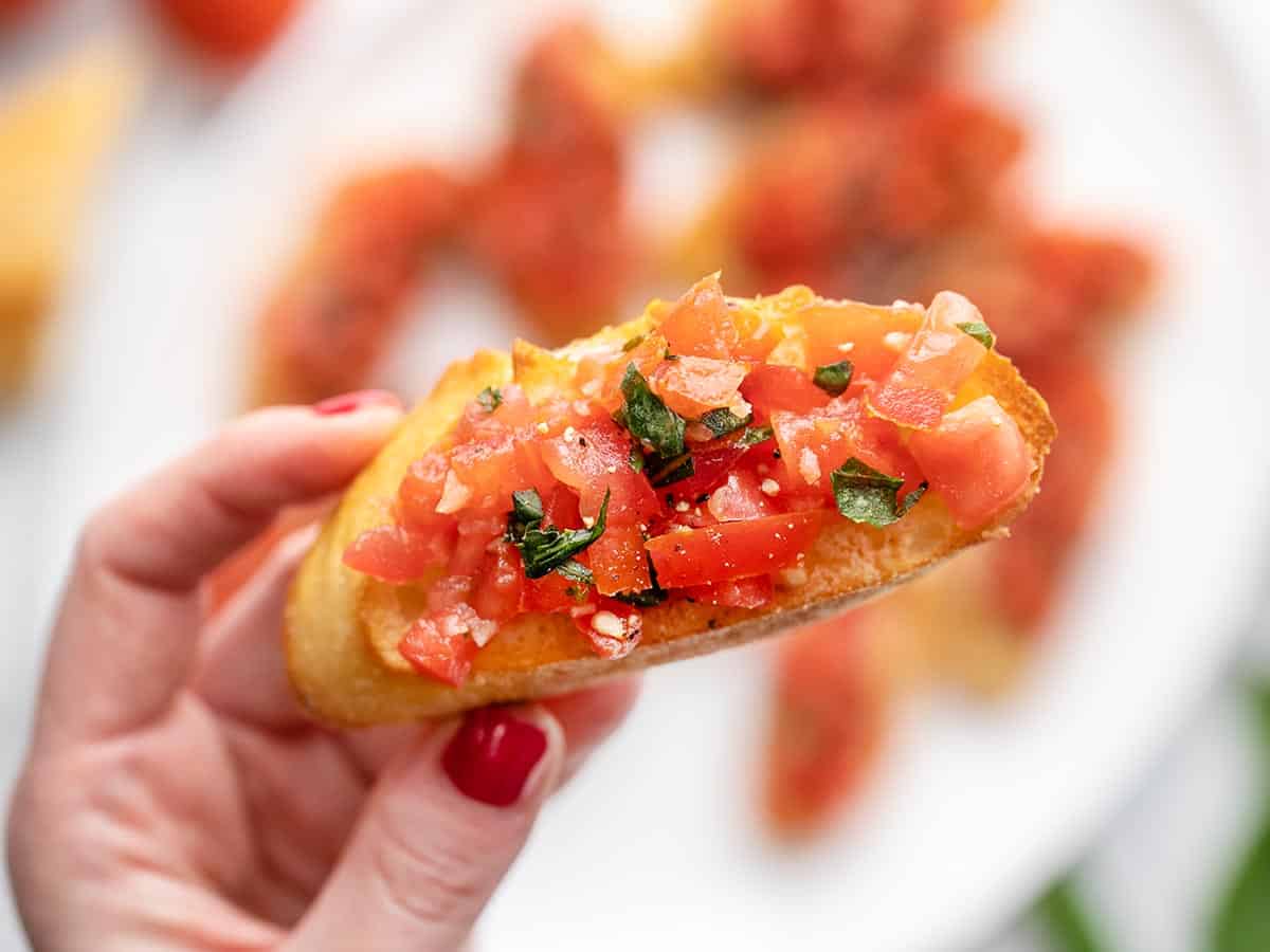 Overhead shot of a hand holding a slice of bruschetta in the foreground and a platter of bruschetta in the background.