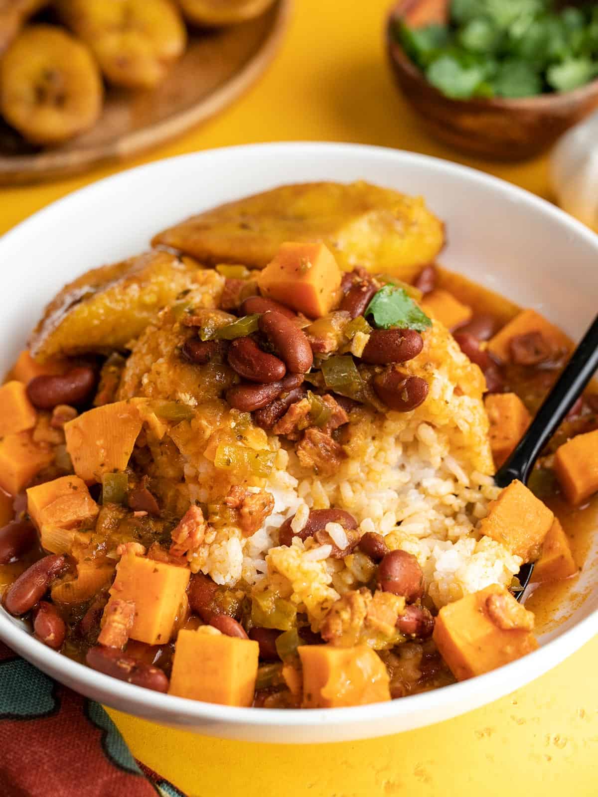 Side shot of a white bowl with red beans and rice and slice ripe plantain with a black spoon in it.