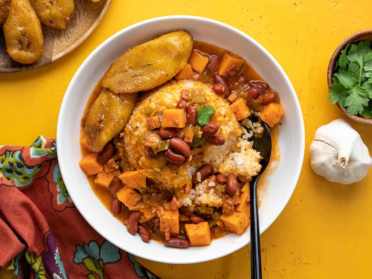 Overhead shot of a white bowl of red beans and rice with a side of ripe plantain slices. and a black spoon in it