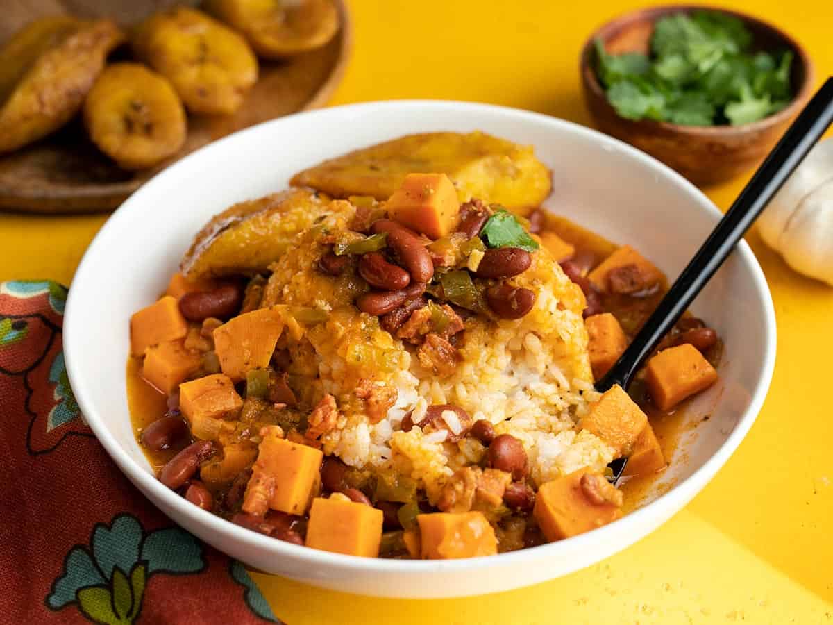 Side shot of a white bowl of red beans and rice with a side of ripe plantains and a spoon in it.