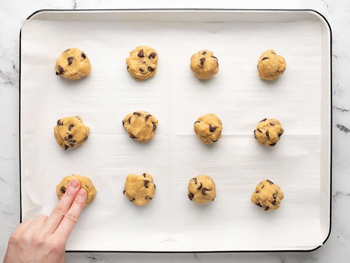 Overhead shot of two fingers pressing down on refrigerated balls of cookie dough on a lined sheet pan.