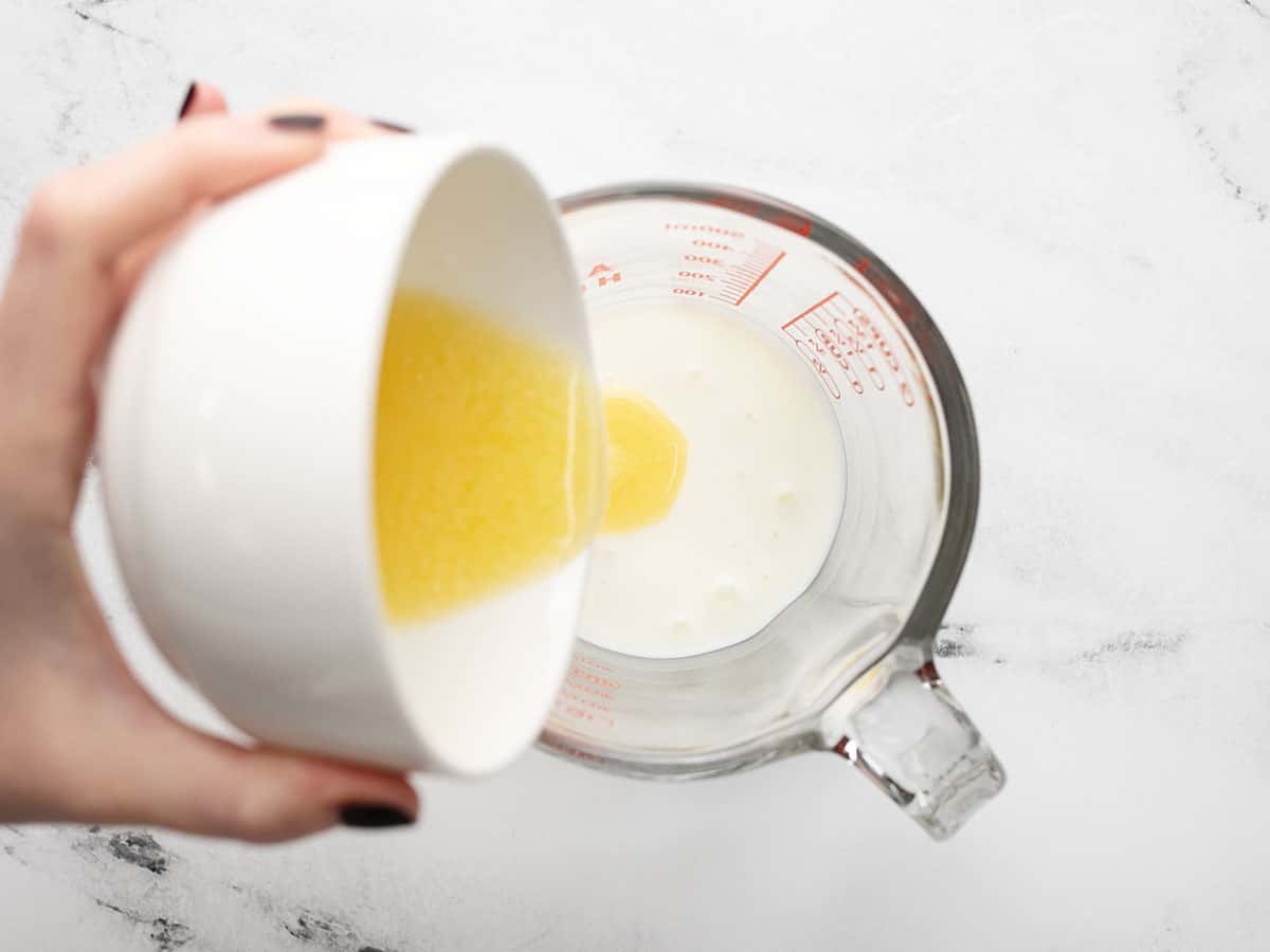 Overhead shot of cook pouring melted butter into a glass measuring cup containing some buttermilk.