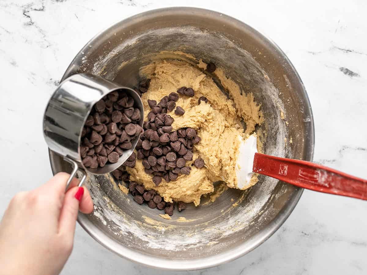 Overhead shot of chocolate chips being mixed into cookie dough with a red and white rubber spatula in a silver bowl.