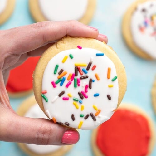 Hand holding a white sugar cookie with sprinkles in the foreground with red and white sugar cookies in the background.