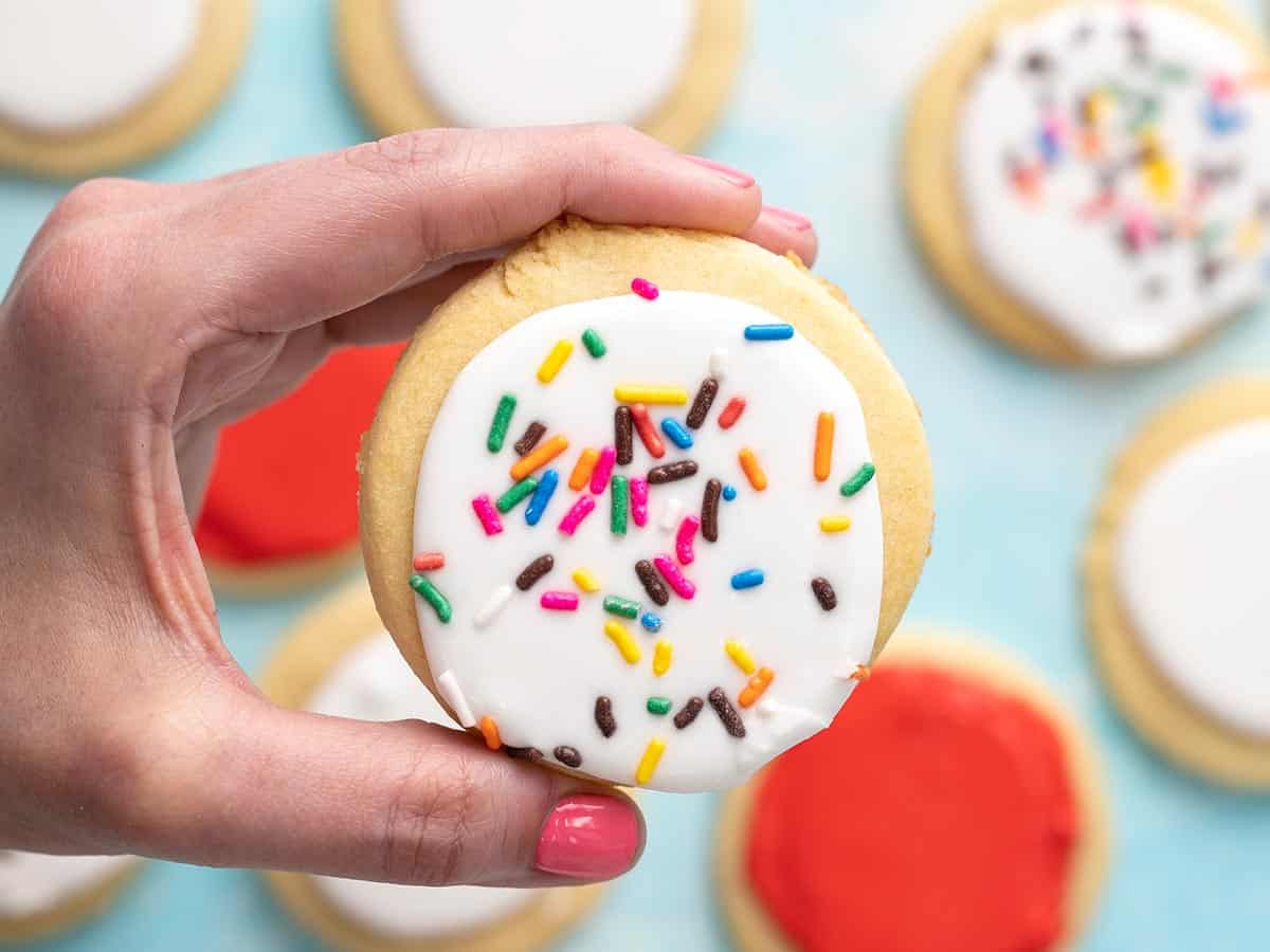 Hand holding a white sugar cookie with sprinkles in the foreground with red and white sugar cookies in the background.