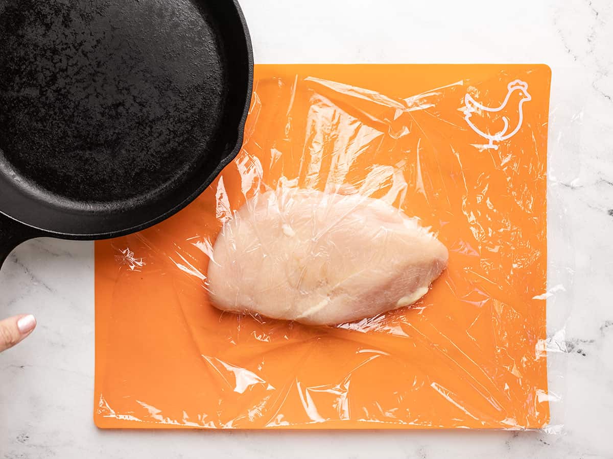 Overhead shot of a chicken breast covered in plastic wrap ready to be flattened by a cast iron pan.