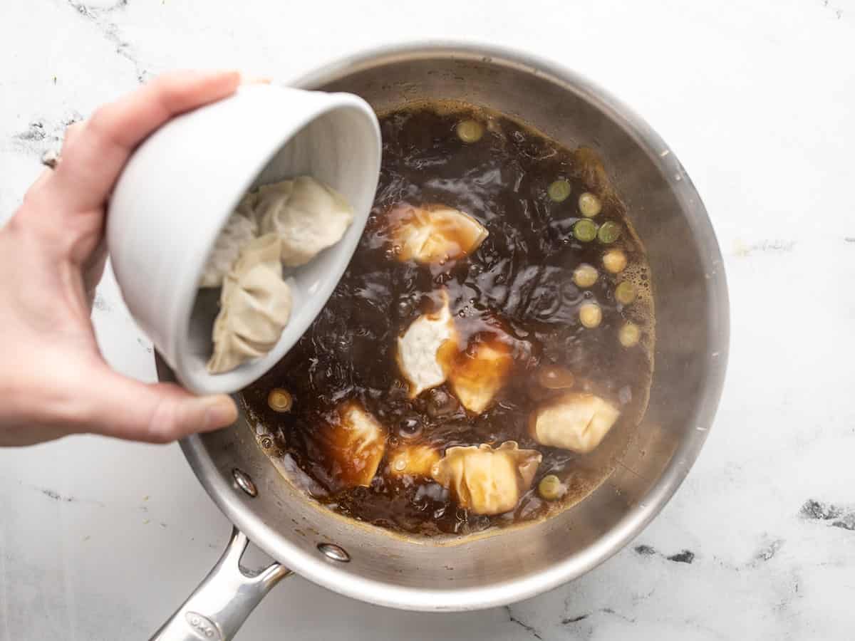 Frozen dumplings in a white bowl being poured into a pot of boiling broth.
