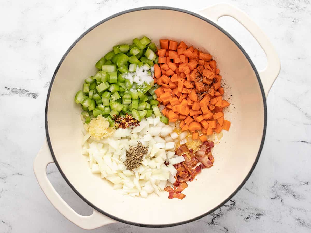 A large dutch oven filled halfway with uncooked diced onions, diced carrots, diced celery with cooked bacon slices underneath and piles of dried red pepper, minced garlic and dried oregano, salt and pepper on top.