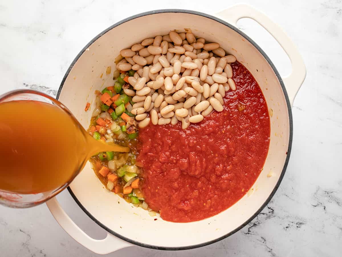 A large pot half filled with sauteed vegetables, uncooked white beans and crushed tomatoes, and a hand overhead pouring vegetable broth into the pot with a liquid measuring cup.
