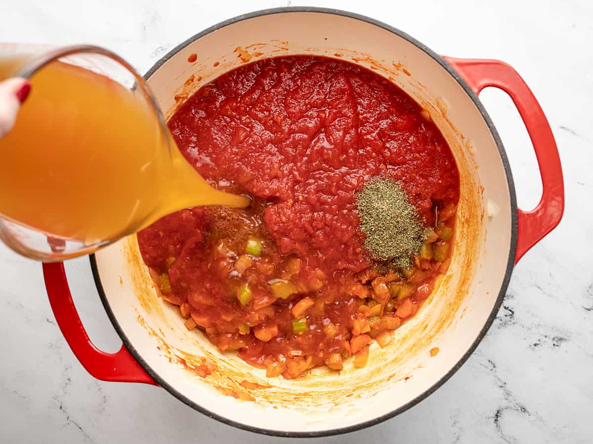 Crushed tomatoes and herbs in the pot, broth pouring in the side. 