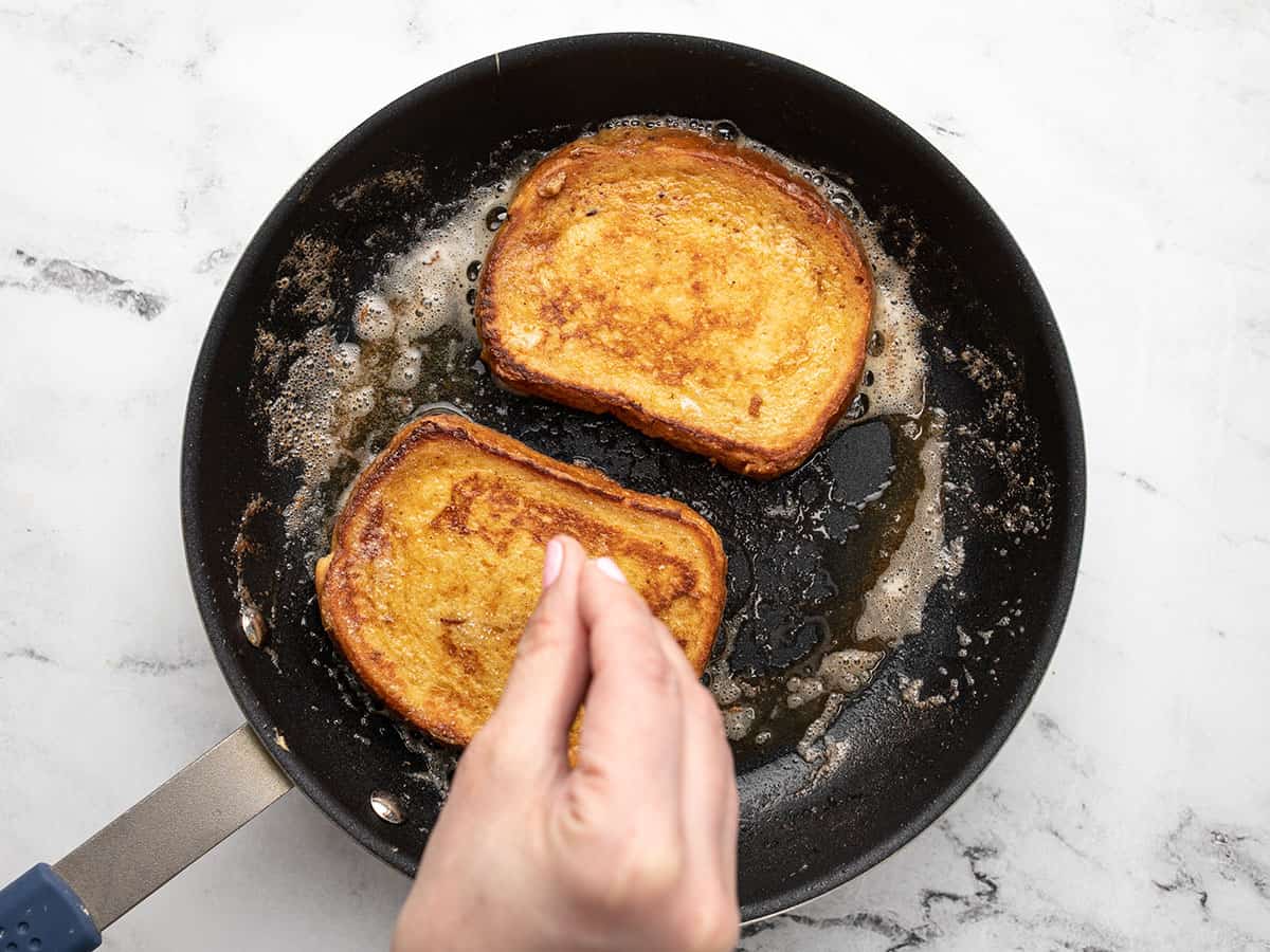 A hand sprinkling sugar over two slices of french toast cooking in a pan.