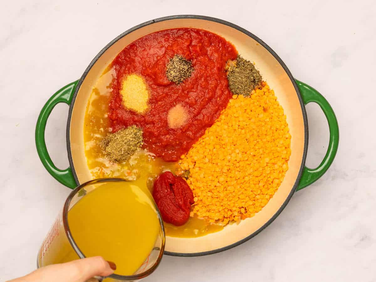 Lentils, tomatoes, and spices in the pot, broth being poured in the side. 