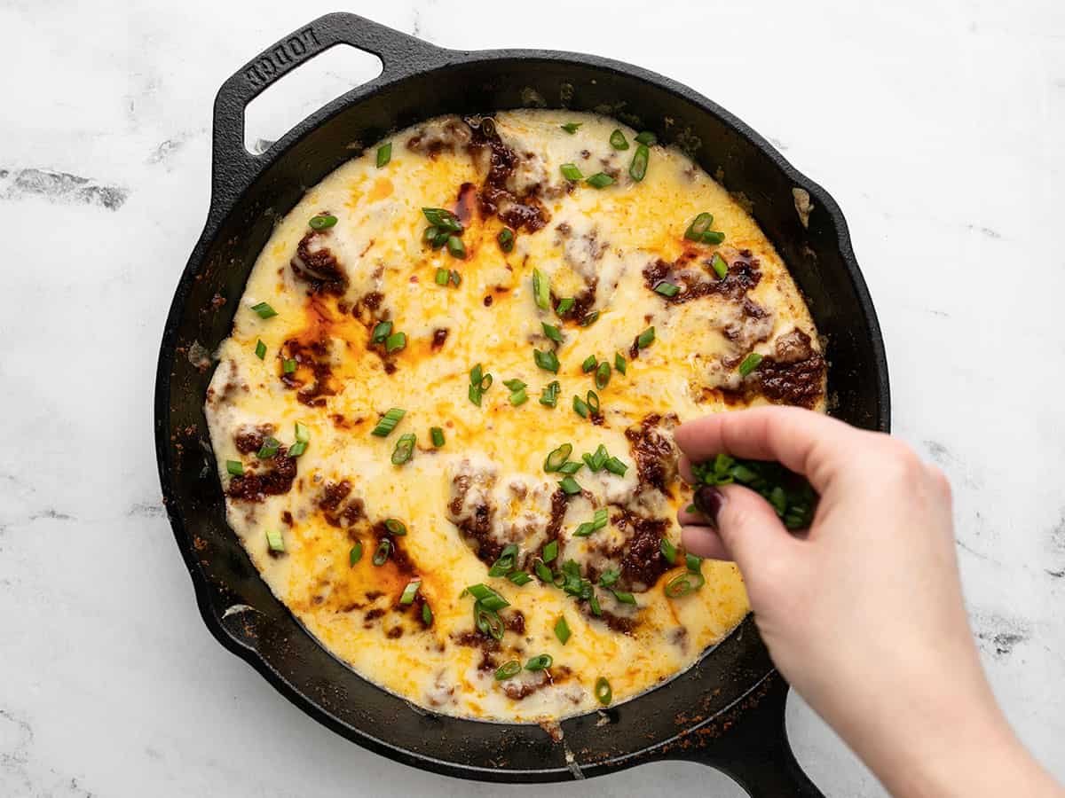Overhead shot of Queso Fundido in a cast iron pan with a hand sprinkling sliced green onions on top.