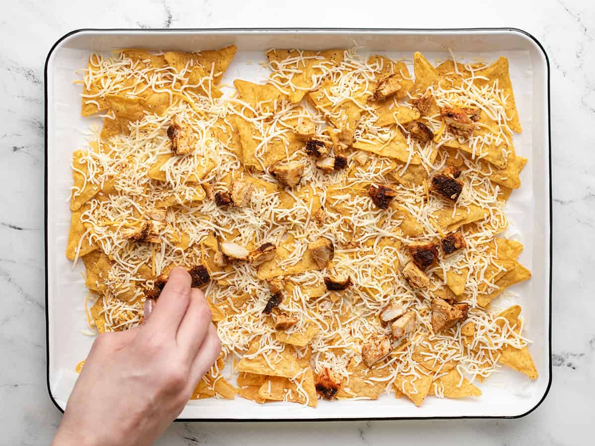 Overhead shot of a hand putting chicken on the first layer of chicken nachos on a sheet pan..