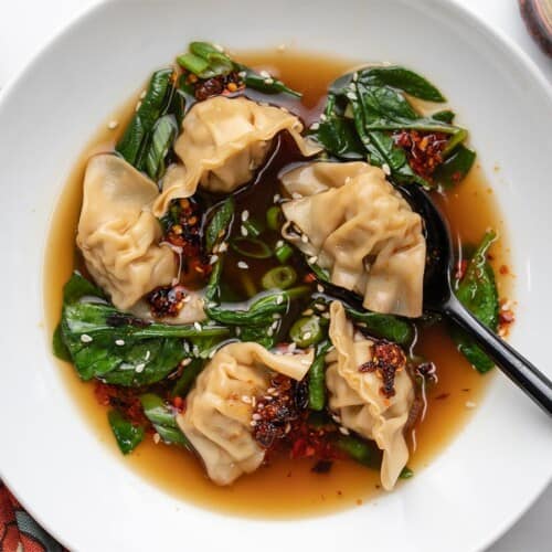 A close up view of dumpling soup in a white bowl topped with white sesame seeds and chili crisp.