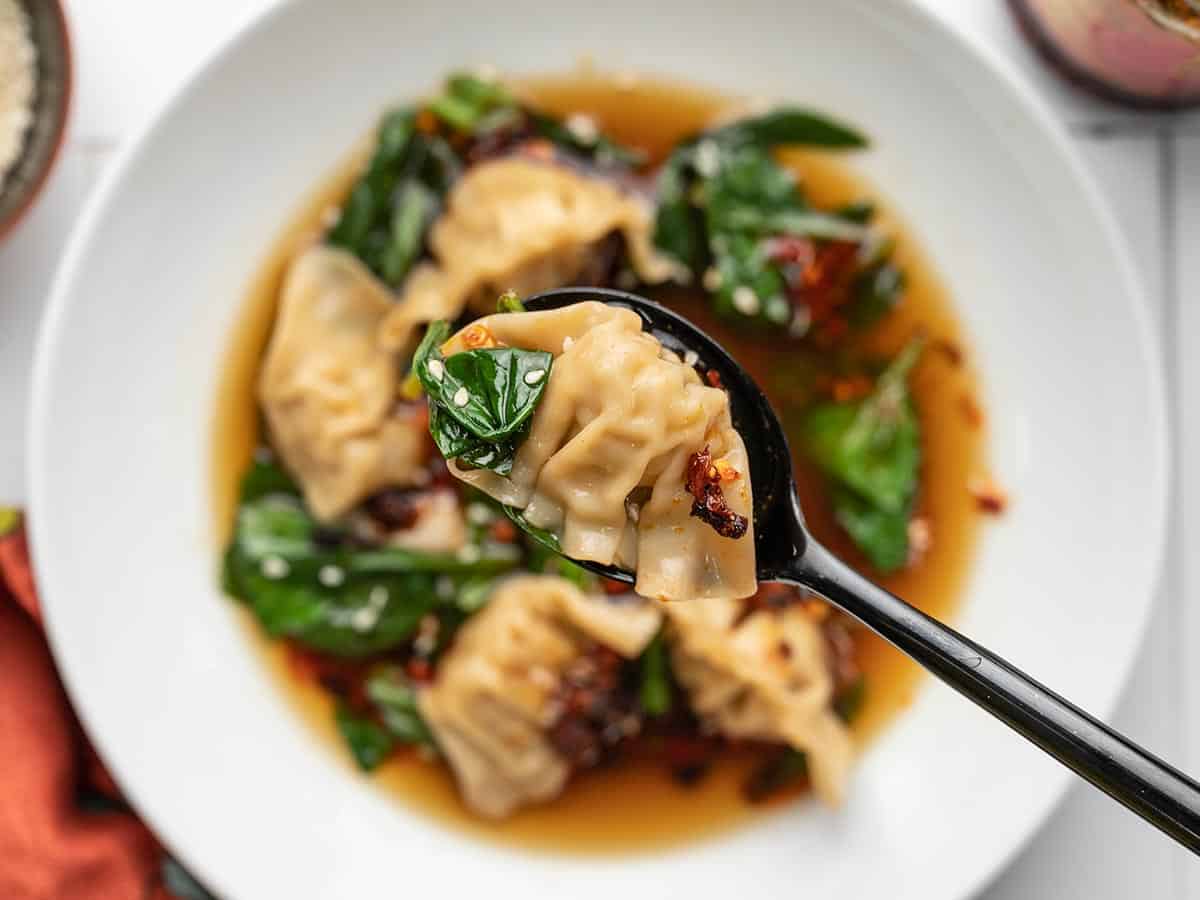 A single dumpling parcel on a black spoon raised above a large white bowl filled with dumpling soup topped with white sesame seeds and red chili crisp and an orange decorative towel.