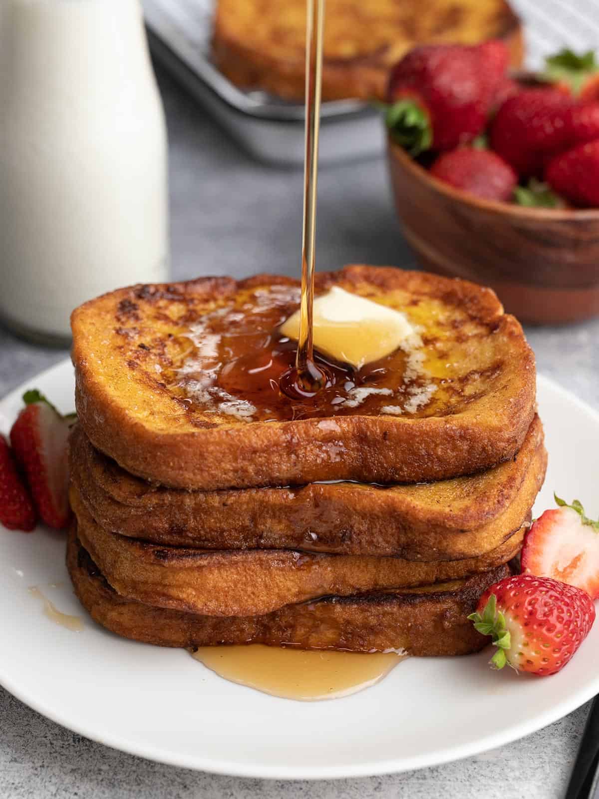 Side view of four slices of french toast stacked on a white plate being drizzled with maple syrup. 