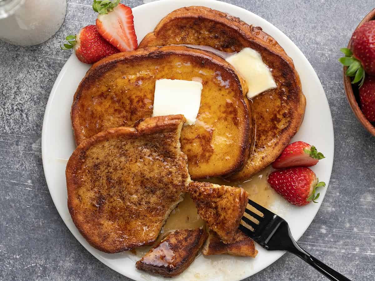 Overhead shot of three slices of cooked french toast on a white plate with sliced strawberries.