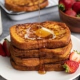 Side view of four slices of french toast stacked on a white plate being drizzled with maple syrup.