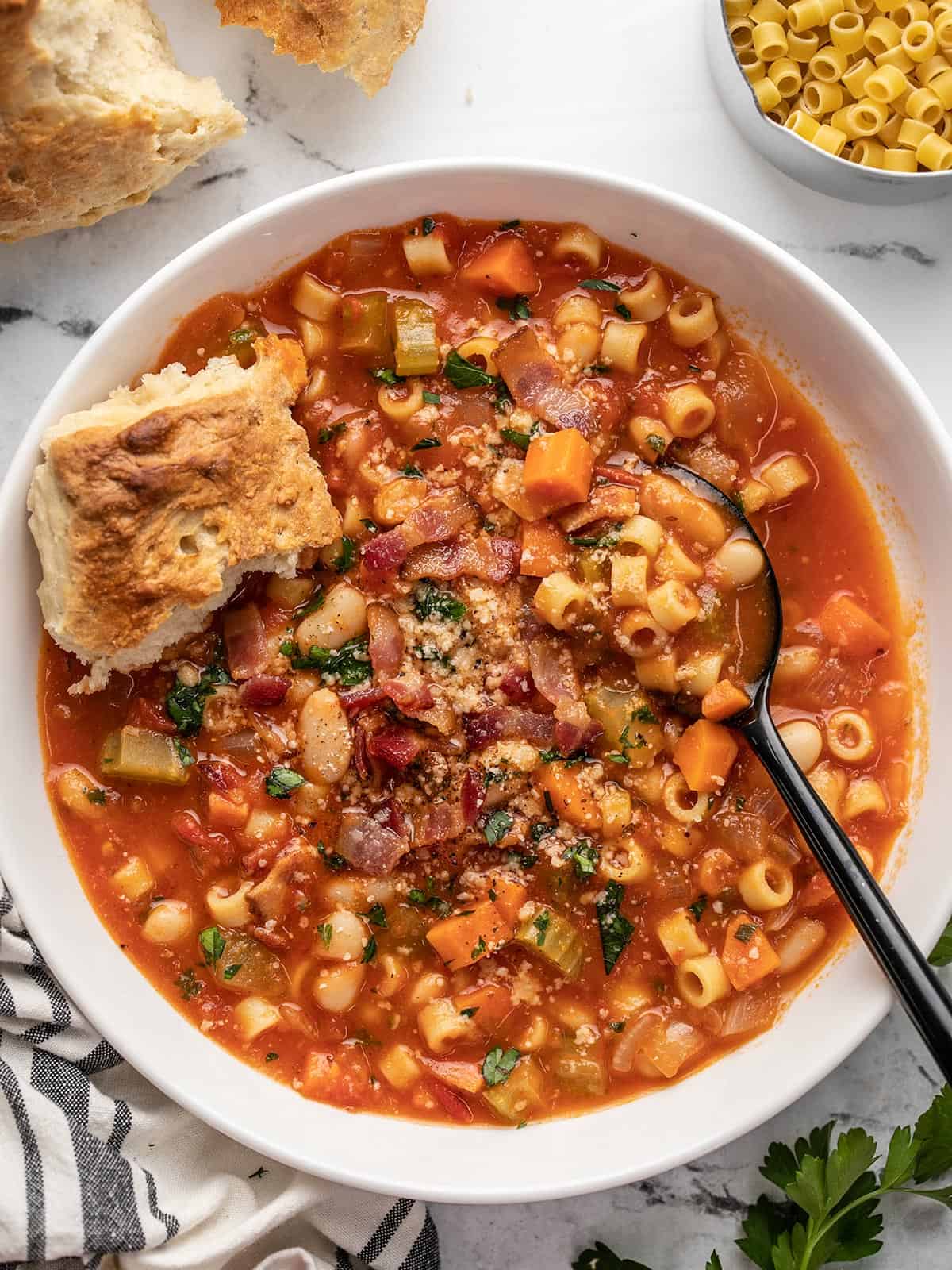 Large bowl of pasta e fagioli soup in a white bowl with a black spoon and topped with a torn piece of bread and surrounded by other ingredients like more bread, uncooked pasta, and a decorative blue and white napkin.