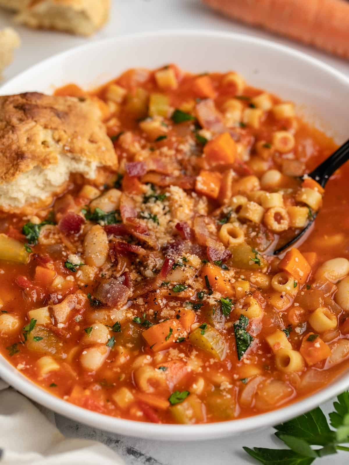 Side view of a bowl of pasta e fagioli soup topped with bacon, parsley and parmesan cheese with a black soup spoon and a torn piece of bread on the side.