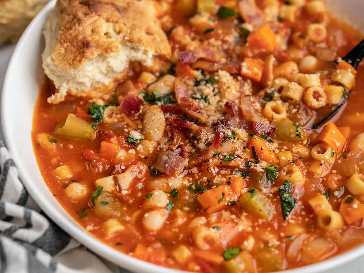 Side view of a white serving bowl filled with Pasta e Fagioli topped with parsley, parmesean cheese, bacon crumbles and crusty Irish Soda Bread.