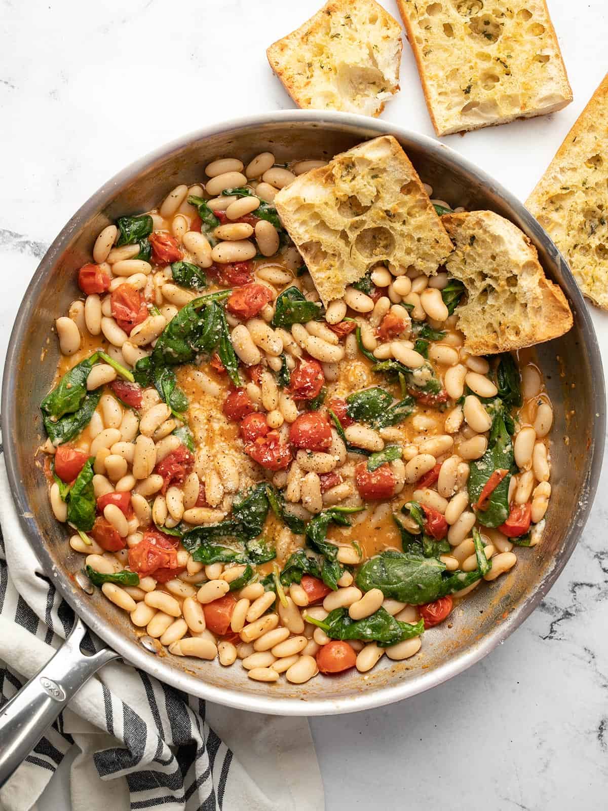 Overhead view of a skillet full of saucy white beans with garlic bread on the side.