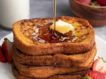 Side view of four slices of french toast stacked on a white plate being drizzled with maple syrup.