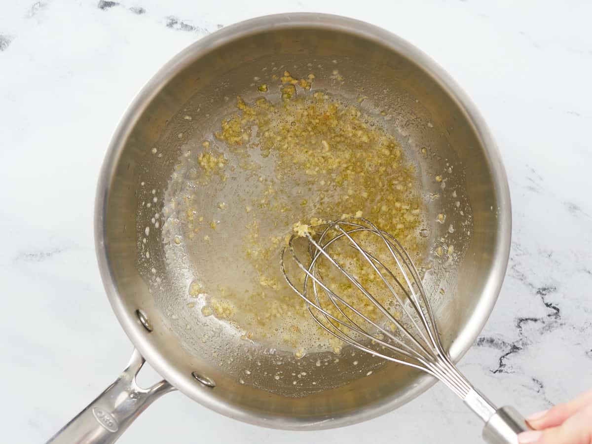 Garlic being sautéed in melted butter and stirred with a balloon whisk in a stainless steel skillet on a white marble backdrop. 